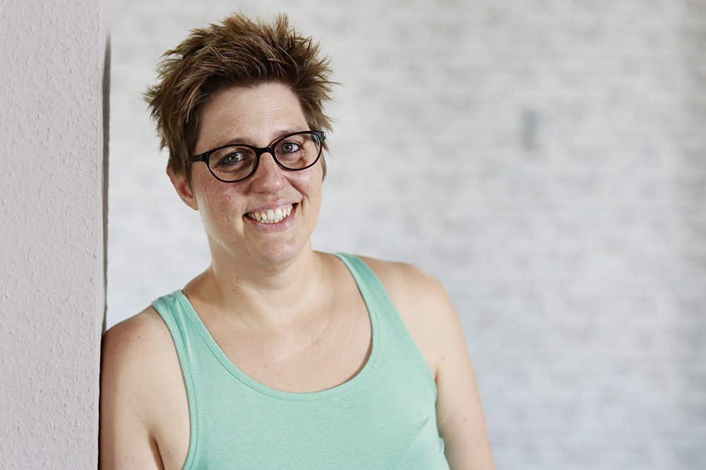 Eine Person mit kurzen Haaren, Brille und einem grünen Tanktop lächelt vor einem weißen Hintergrund.